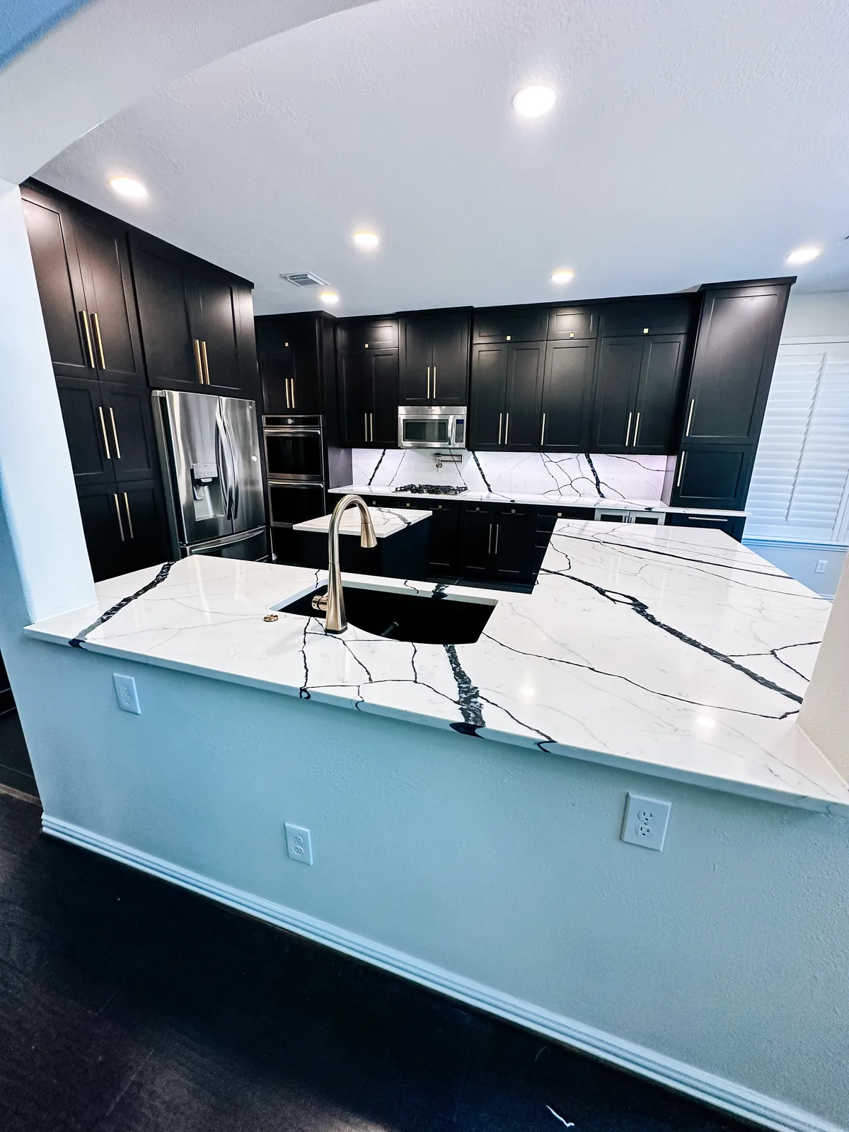 Open-concept remodeled kitchen with dark shaker cabinets, black-veined quartz peninsula with undermount sink and brass faucet, and stainless appliances