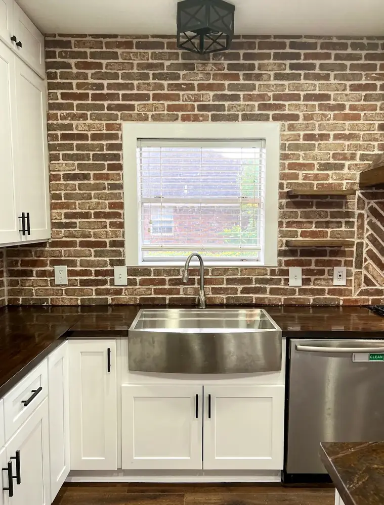 Stainless farmhouse apron sink set into dark butcher-block counters beneath a window in an exposed-brick kitchen
